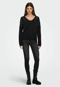 Pull noir à col en V avec des sequins sur les épaules, associé à un jean noir slim et des bottines. L'ensemble est monochrome.