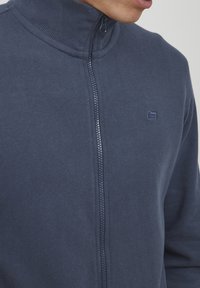 Sudadera navy con cremallera y cuello alto, con textura acanalada y detalle de logo. Tejido suave con cierre de cremallera lisa.