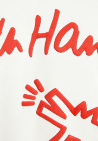 Röd grafisk text och design på ett vitt tyg, med stora bokstäver och en tydlig handsymbol med strålande linjer. Mjuk textur.