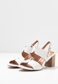 Sandali bianchi con tacco alto, design a ritaglio, cinturini in pelle e tacco a blocco marrone. Presentano una cinghia alla caviglia regolabile e una texture liscia.