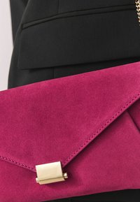 Pochette enveloppe en daim rose vif avec fermoir doré tenue contre un vêtement en tissu noir.