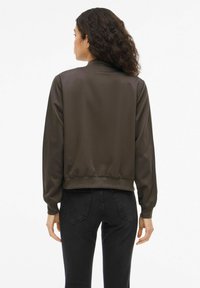 Brun bomberjakke med glatt, skinnende tekstur og ribbestrikkede mansjetter, båret over svarte jeans. Enkel design uten pynt.