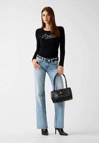 Haut noir à manches longues avec logo argenté, associé à un jean évasé délavé clair et un sac à main noir. Mannequin portant des bottes noires à talons hauts.