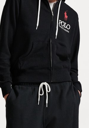 Person trägt schwarzen Kapuzenpullover mit Reißverschluss, weißen Kordeln und Polo Ralph Lauren Logo in Rot und Weiß, kombiniert mit schwarzen Hosen mit weißen Kordeln.