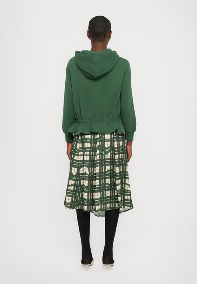 Felpa verde con vita cinchata e dettaglio peplum, abbinata a una gonna a quadri crema con righe verdi; collant neri e décolleté a punta.