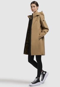 Lange, tan regenjas met capuchon, ritsluiting en zwart interieurpatroon. Gecombineerd met zwarte leggings en hoge sneakers.