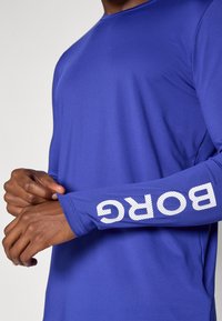Ilgos rankovės sportinė marškinėliai ryškiai mėlynos spalvos audinyje, pasižymintys glotnia tekstūra ir balta "BORG" spauda prie riešo.