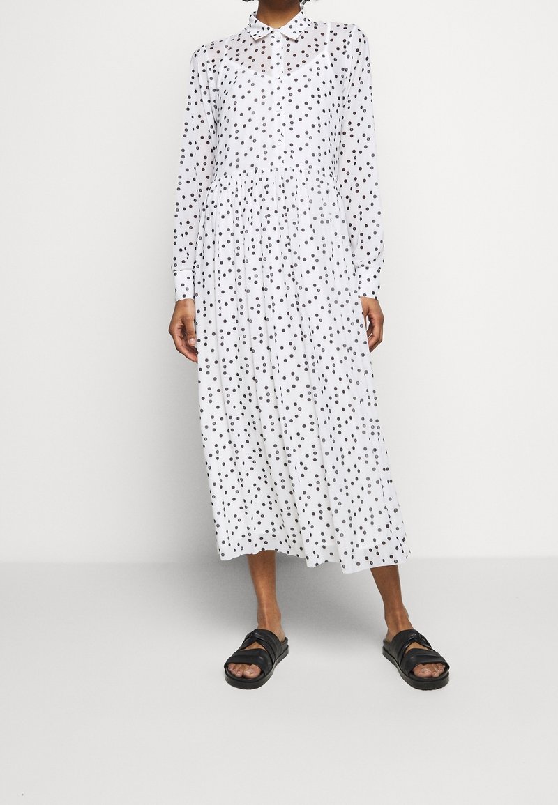 Personne portant une robe midi blanche à manches longues avec des pois noirs et des sandales noires à bout ouvert, debout devant un fond clair uni.
