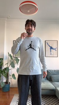 Pikkade varrukatega hall särk, mille ees on must Jordan logo, millele on lisatud tumesinised püksid. Siseruum, kus on taimi ja mööblit.