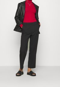 Blazer en cuir noir, col roulé rouge côtelé, pantalons noirs ajustés et sandales à glissière noires. Présente une coupe décontractée et un design minimaliste.
