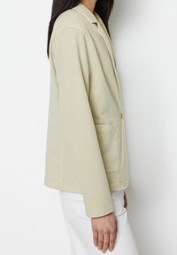 Jasny żółty żakiet o miękkiej teksturze, z klapami z wycięciem, pojedynczym zapięciem na guzik i kieszeniami po bokach. Długie rękawy, luźny krój.