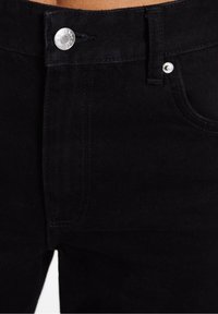 Jeans de mezclilla negra con cierre de botón estándar, bolsillos delanteros y detalles de costura visibles. El material es suave con un ligero brillo.