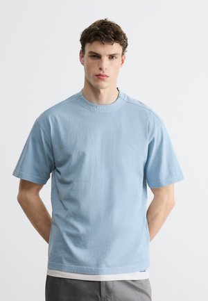 Giovane uomo con capelli castani corti indossa una maglietta azzurra chiara sopra una canottiera bianca, in piedi di fronte a uno sfondo chiaro uniforme.