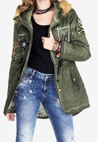 Parka vert olive avec col en fausse fourrure, ornée de patchs et de décorations ; associée à un jean bleu déchiré avec des clous en argent.