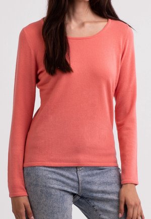 Kvinde iført en koralfarvet langærmet sweater og lyseblå jeans, stående med arme afslappet ved siden.