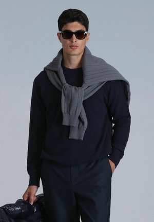 Pull en marine, écharpe en tricot grise drapée sur les épaules, lunettes de soleil noires, tenant une doudoune noire brillante. Textures lisses, design contemporain.
