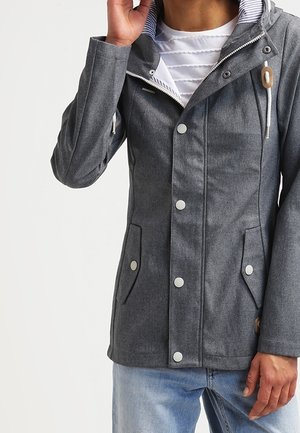 Homme portant une veste à capuche grise avec des fermetures à boutons par-dessus une chemise blanche texturée et un jean bleu clair, touchant la capuche.