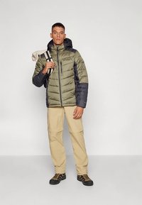 Moški v zeleni in črni izolirani jakni, bež hlačah, pohodniških čevljih, nosi zavit bež mat čez ramo na belem ozadju.