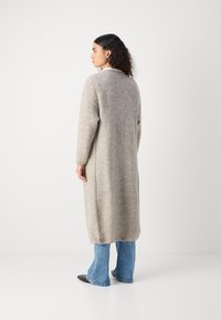 Long, grey knitted cardigan with full sleeves and side slits, paired with flared blue jeans and black pointed shoes. Smooth, soft texture.