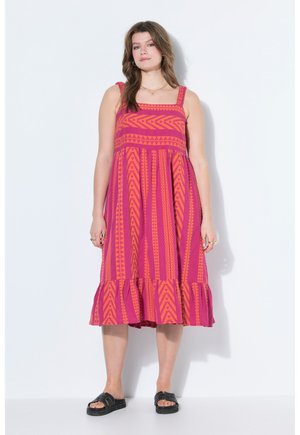 Kobieta w różowo-pomarańczowej sukience midi bez rękawów z falbanką u dołu i czarnych sandałach na wsuwkę, stojąca na białym tle.