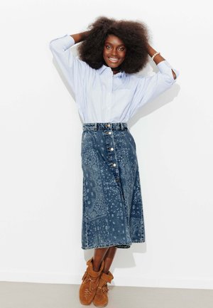 Femme souriante aux grands cheveux bouclés portant une chemise rayée bleue, une jupe en jean à motifs et des bottines mocassins à franges marron, posant contre un mur blanc.