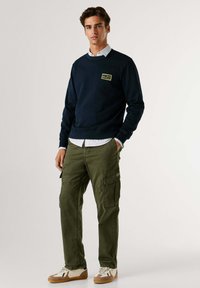 Marineblauwe sweatshirt met een geel logopatch, gecombineerd met olijfgroene cargobroek. De outfit heeft een casual ontwerp en een ontspannen pasvorm.
