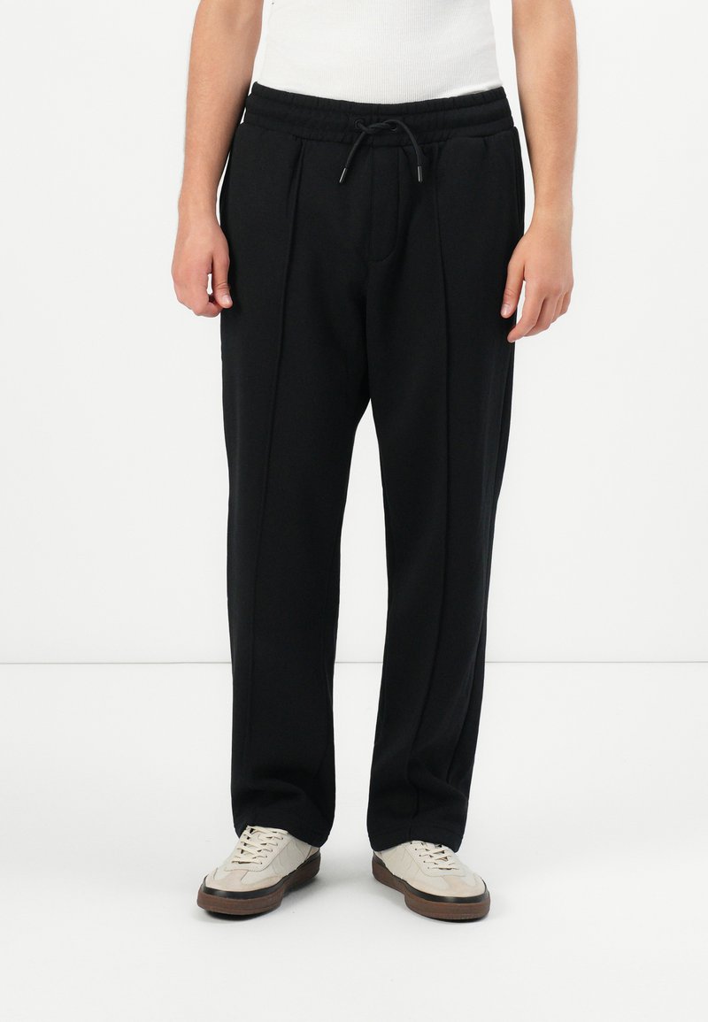 Persona vestida con pantalones rectos negros con una cinturilla elástica y zapatillas blancas, de pie contra un fondo blanco.