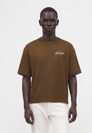 Ung mand med mørk hud iført brun Polo Sport t-shirt og hvide bukser står foran en ensfarvet hvid baggrund.