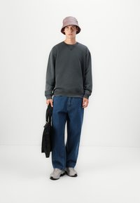Sudadera gris con hombros caídos, jeans anchos azules, zapatillas grises y un sombrero tipo cubo rosa claro. Sosteniendo un bolso negro con una correa.