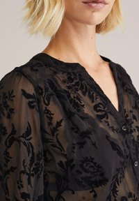 Camicia nera trasparente con motivi floreali ricamati, colletto e chiusura con bottoni, realizzata in tessuto morbido e testurizzato.