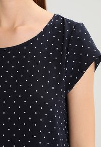 Námořnická modrá blůza s krátkými rukávy a kulatým výstřihem, s bílým puntíkatým vzorem. Měkká, lehká tkanina, hladká textura.