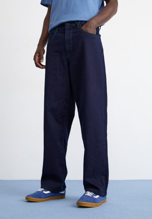 Marineblå viddejeans i et robust stof med kontrasterende grønne syninger og sidelommer, parret med blå sneakers og en lyseblå skjorte.