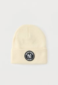 Căciulă crem din material tricotat moale. Are o emblema rotundă neagră cu litere albe și o margine pliată la bază.