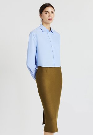 Frau in einem hellblauen kurzärmeligen Hemd und einem hochgeschnittenen olivgrünen Midikleid steht vor einem schlichten Hintergrund.
