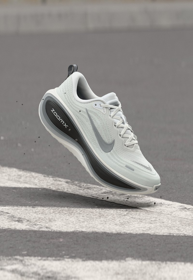 Λευκό και γκρι παπούτσι τρεξίματος Nike ZoomX να αιωρείται πάνω από μια σηματοδοτημένη ασφαλτοστρωμένη επιφάνεια με σωματίδια βρωμιάς γύρω από την σόλα.