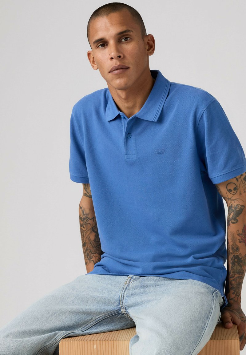 Joven con el cabello corto, camiseta polo azul y vaqueros claros sentado sobre un bloque de madera con un fondo liso.