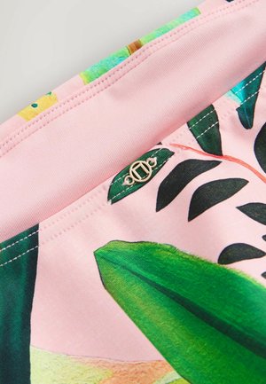 Roze stof met groene bladprint en gedetailleerd stikwerk, voorzien van een klein rond metalen logo-embleem genaaid bij een naad.