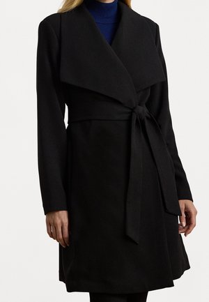 Femme portant un manteau noir mi-long avec de larges revers et une ceinture nouée à la taille, par-dessus un pull à col roulé bleu.