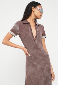 Žena v hnedom krátkom rukáve šatách s hlbokým výstrihom do V a gombíkmi, pózujúca s rukou na bokoch, nosiaca veľké reflexné slnečné okuliare.