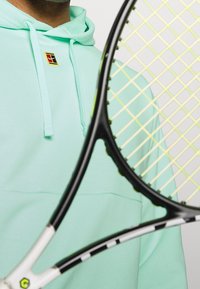Sweat à capuche vert menthe avec cordons de serrage, patch logo, et une raquette de tennis noire avec un motif de cordes jaunes, partiellement visible.