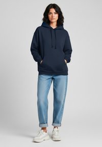 Felpa blu navy con tasca frontale, cordoncini e vestibilità rilassata, abbinata a jeans azzurri arrotolati e sneakers bianche.