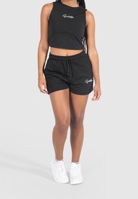 Donna che indossa un top corto senza maniche nero e pantaloni corti coordinati, con sneakers bianche, in piedi davanti a uno sfondo chiaro e uniforme.