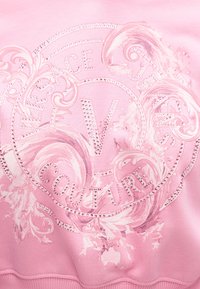 Material roz cu logo-ul Versace Jeans Couture într-un design circular și cu aplicații cu ținte care înconjoară modele decorative în stil baroc.