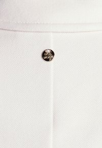 Detail bílého texturovaného látkového materiálu s malým kulatým kovovým knoflíkem označeným "MOS MOSH", umístěným na svislém švu.