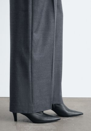 Bottines noires à bout pointu portées avec un pantalon gris long à jambes larges, debout sur un sol en béton poli.