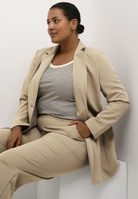 Costume beige ajusté avec un blazer à simple boutonnage, haut rayé noir et blanc, tissu lisse et coupe décontractée, assis contre un fond clair.