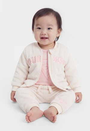 Tout-petit souriant assis pieds nus, portant une veste et un pantalon rose clair avec le logo "Guess" sur un fond blanc uni.