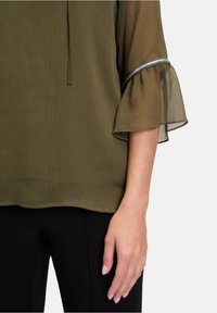 Blouse vert olive en tissu texturé, manches évasées transparentes et bordure d'accent argentée. Associée à un pantalon noir, montrant une main posée.