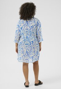 Blå och vit mönstrad kimono-liknande cover-up med lös passform, ärmar i medellängd och en lätt, genomskinlig tyg. Platta sandaler bärs.