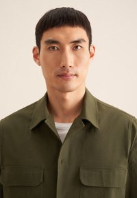 Chemise olive en tissu léger avec un col évasé, dotée de deux poches avant, en coton, et d'une texture lisse.